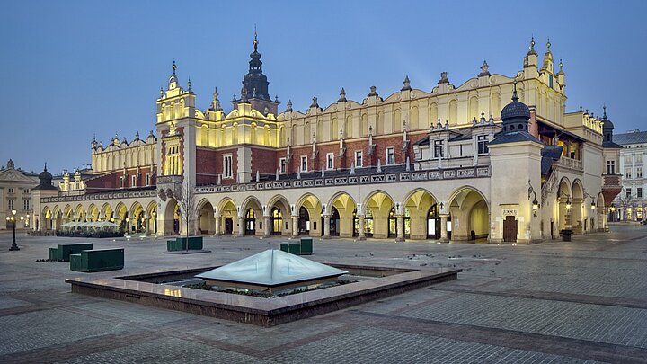 Zgjęcie Głównej Płyty Rynku w Krakowie, na pierwszym planie fontanna "Kryształ", na dalszym planie Sukiennice, zza sukiennic wystaje Wieża Ratuszowa